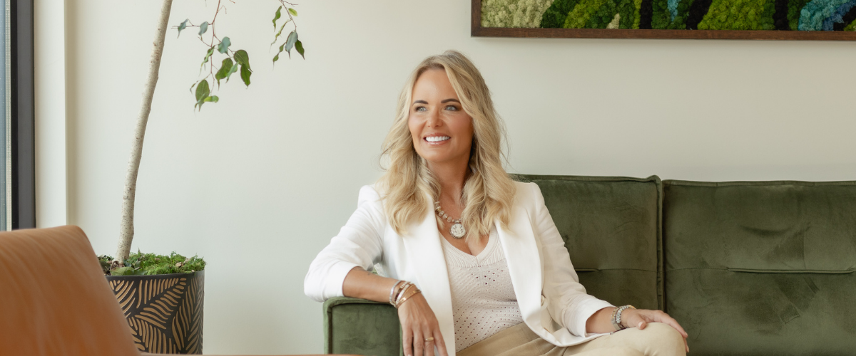 A woman in business attire sitting on a green couch in an office loking out the window and smiling 